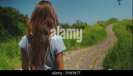 A woman with long hair is walking down a dirt road in Ubud, Bali. The - captures her movement along the Campuhan Ridge Walk. Stock Photo