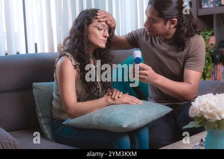 A man measures a woman's temperature with a digital infrared thermometer at home sitting on the couch, The image highlights health and safety protocol Stock Photo