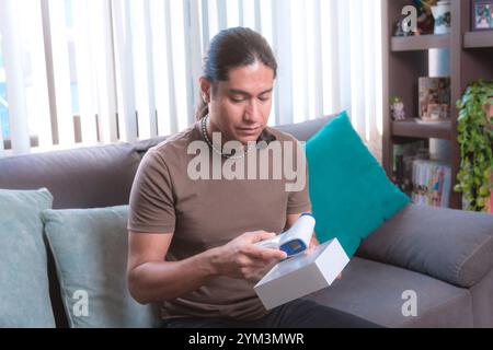 A man measures his temperature with a digital infrared thermometer at home sitting on the couch, The image highlights health and safety protocols in a Stock Photo