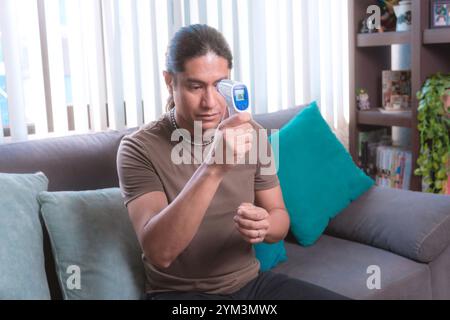 A man measures his temperature with a digital infrared thermometer at home sitting on the couch, The image highlights health and safety protocols in a Stock Photo