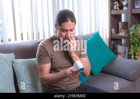 A man measures his temperature with a digital infrared thermometer at home sitting on the couch, The image highlights health and safety protocols in a Stock Photo
