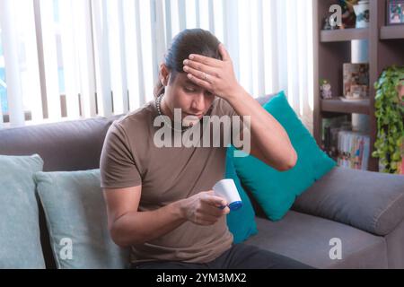 A man measures his temperature with a digital infrared thermometer at home sitting on the couch, The image highlights health and safety protocols in a Stock Photo
