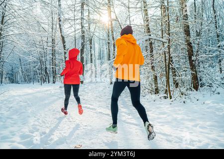 Young couple trail runners man and woman dressed bright windproof jackets running in picturesque snowy forest during sunny frosty day. Sporty active p Stock Photo