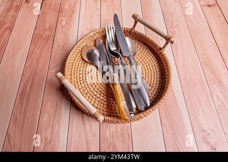 A wicker tray with wooden handles filled with metal cutlery on a honey-colored wooden table Stock Photo