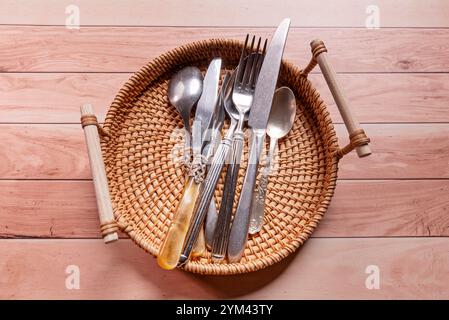 Overhead view of a wicker tray with wooden handles filled with metal cutlery on a honey-colored wooden table Stock Photo