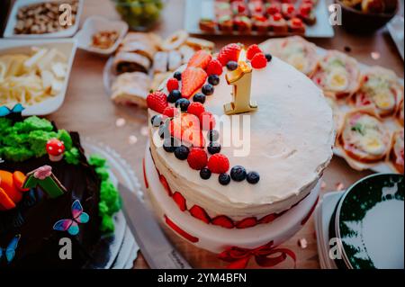 Close-up of a vibrant fruit cake decorated with fresh berries and a '1' cake topper, symbolizing the celebration of a child's first year of life Stock Photo