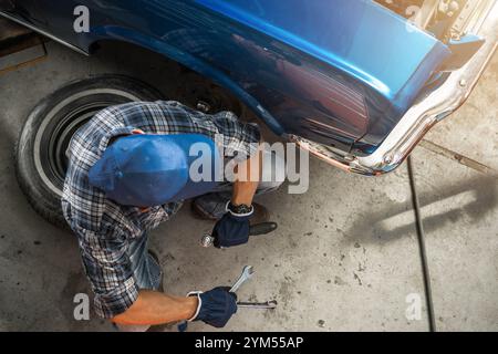 A mechanic focuses on repairing a classic car in a garage, surrounded by tools and an old tire, emphasizing dedication to the craft. Stock Photo