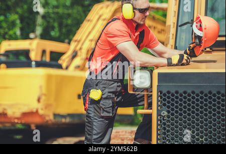 A construction worker in orange gear inspects machinery at a busy site, ensuring safety and proper operation with protective equipment. Stock Photo