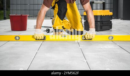A construction worker is using a level tool to ensure tile alignment on a floor at a construction site. The worker is focused and dressed for the task Stock Photo