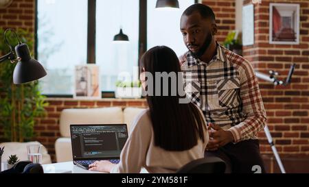 Interracial creative team programing code on a laptop and debugging, using digital tools at home. Professional coders team working on software development and internet security. Camera B. Stock Photo