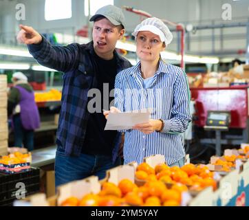 Workers of fruit storage and sorting facility talking about work process standing near the conveyor line and tangerine boxes. Stock Photo