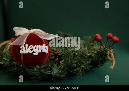 A red ball with Alegria written on it is placed on top of a green wreath. The wreath is made of evergreen branches and has a few pine cones scattered Stock Photo