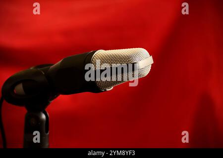 Close-up of a studio microphone against a dark red background, featuring a metal grille and black body, perfect for podcasts or music recording. Stock Photo