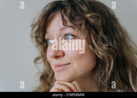 Close-up portrait of a young woman with curly brown hair, resting her chin on her hand, gazing thoughtfully into the distance with a gentle smile, conveying a sense of contemplation and optimism Stock Photo