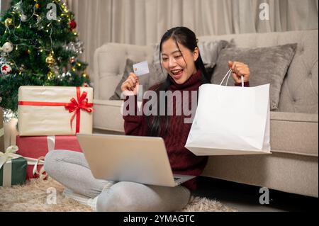 An excited Asian woman in cozy clothes enjoys shopping online at home during the Christmas holiday, pleasantly surprised by the big sales on the shopp Stock Photo
