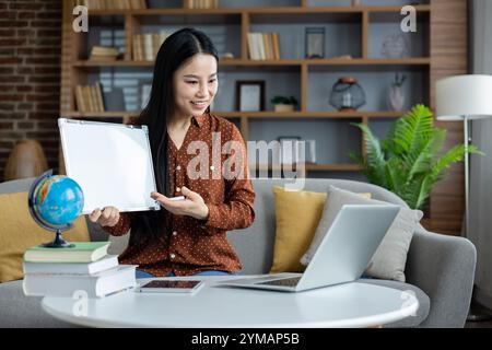 Asian woman conducting an online lesson with laptop and whiteboard. Engaging interaction through digital medium, integrating global and educational concepts from home. Stock Photo