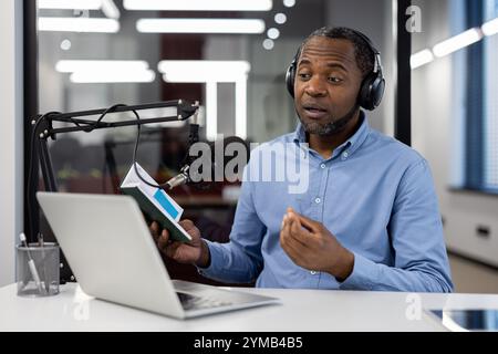 Mature African American businessman recording podcast in modern studio. Wearing headphones, using microphone and laptop, sharing insights passionately. Engaged, focused, professional environment. Stock Photo