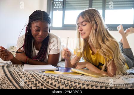 Multiethnic students studying together, lying on bed with notebooks and pens Stock Photo