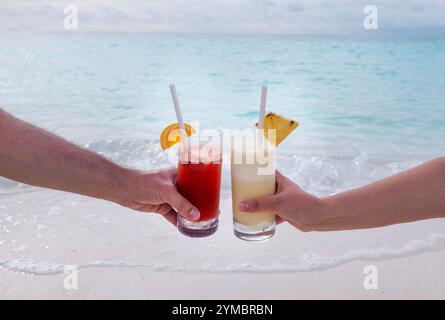 A close-up of a male and a female hands holding two glasses with cocktails. Delicious chilled drinks on the background of the sea and the beach. Young Stock Photo