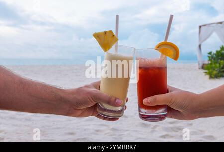 A close-up of a male and a female hands holding two glasses with cocktails. Delicious chilled drinks on the background of the sea and the beach. Young Stock Photo