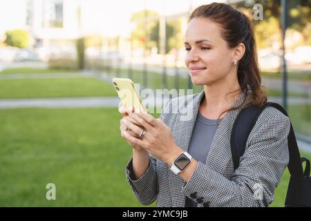 Modern urban woman in smart casual clothes texting online on her smartphone while standing outdoors on a city street. Stock Photo