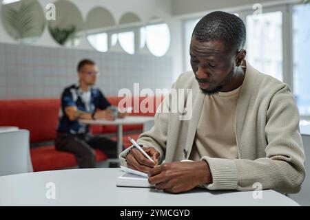 Medium shot of concentrated African American businessman writing in notebook making notes while working or studying at table in sunlit cozy coworking Stock Photo