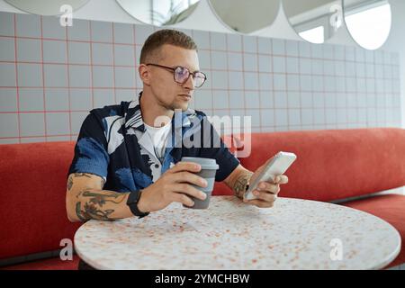 Medium shot of young concentrated man with glasses looking at phone screen checking messages over cup of coffee while working at cafe table, copy spac Stock Photo