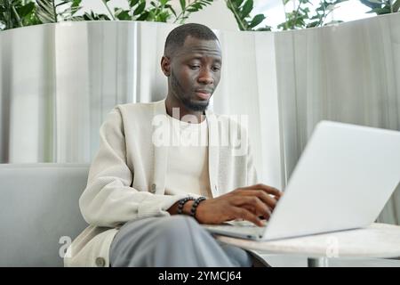 Medium shot of concentrated African American businessman typing on laptop keyboard while working in lounge zone of contemporary coworking space with b Stock Photo