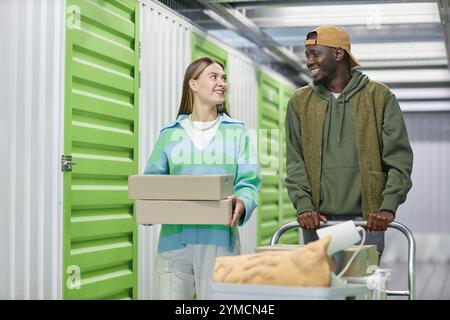 Medium shot of cheerful woman carrying cardboard boxes walking along self storage warehouse hallway with helping boyfriend of African American ethnici Stock Photo