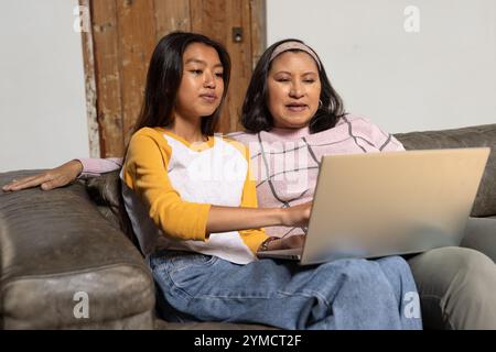 Grandmother and asian female teenager using laptop together on couch at home, sharing moments Stock Photo