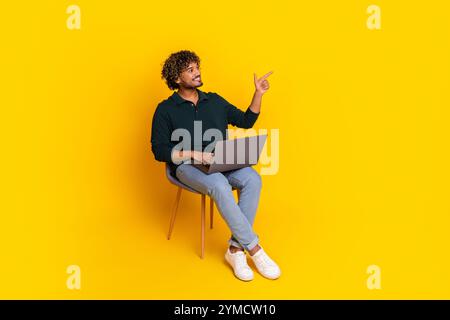 Young man in casual attire using laptop while pointing up with curiosity on vibrant yellow background. Stock Photo