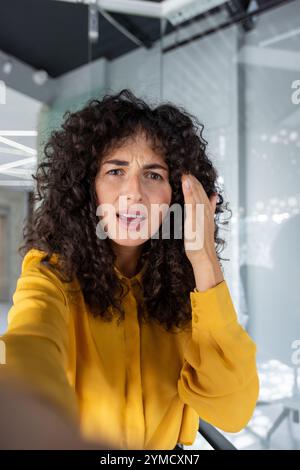 Curly-haired woman making video call using phone, expressing concern or confusion in modern office setting. Bright yellow shirt adds contrast to scene. Emphasizes emotions during call. Stock Photo