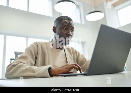 Medium shot of concentrated African American businessman typing on laptop while working in coworking space featuring light interior, copy space Stock Photo