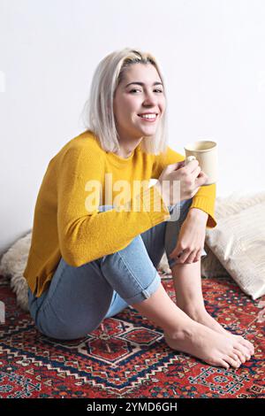 Caucasian woman with blonde hair, in a yellow sweater and jeans, smiling warmly while holding a mug. Relaxed and cozy atmosphere on a patterned rug. Stock Photo