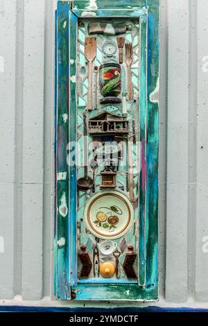 Kitchen utensils – a wooden spoon, a fork, a ladle, a hand-cranked coffee grinder, a painted ceramic bowl with a parrot motif and a dessert plate – fill a long wooden frame of the wall installation 'Memora di Kaya' by TOPO 13, set against a turquoise mosaic background. The installation hangs on a wall next to the Aruba Viaduct, in the Otrabanda district of Willemstad, Curaçao. Stock Photo