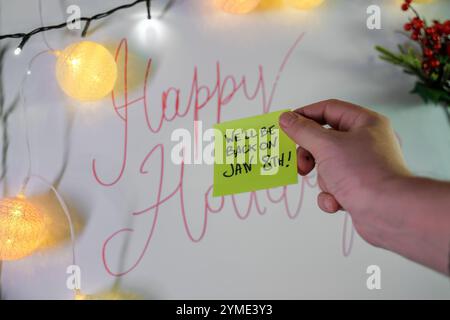 Hand holding yellow sticky note with return date message against white wall with holiday lights and festive decorations Stock Photo