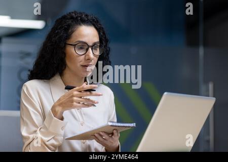 Hispanic business woman participates in online meeting using laptop, taking notes in modern office with glass walls. She exemplifies professionalism and effective communication. Stock Photo