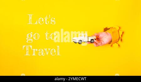Person is holding a toy truck and the words Let's go to travel are written below them. The image has a yellow background and the words are in white Stock Photo