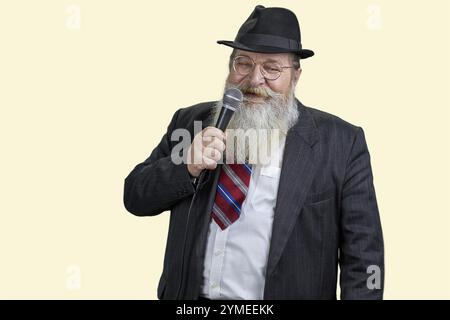 Portrait of old bearded man singing with microphone. Isolated on pastel yellow background Stock Photo