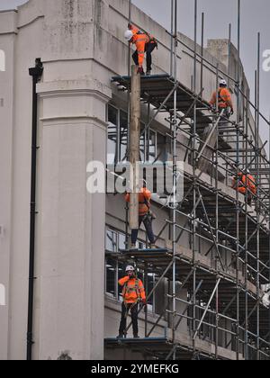 Scaffold being erected by a team of scaffolders in orange hi-vis and hard hats working together to lift scaffold boards to the top platform Stock Photo