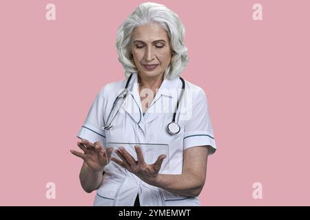 Female doctor with stethoscope using futuristic transparent tablet pc. Senior woman doctor working on glass digital tablet on pink background Stock Photo