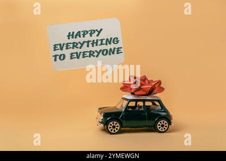 Small green car with a red bow on top of it. The car is surrounded by a speech bubble that says Happy everything to everyone. Concept of joy and celeb Stock Photo