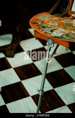 An artist's paint palette with vibrant colors, resting on an easel, set against a black and white checkered floor, with artist's chair Stock Photo