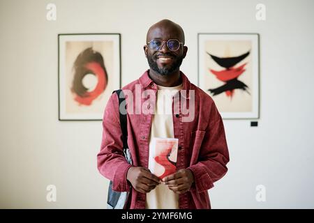 Smiling Black person dressed in casual attire holding red book while standing in art gallery. Two framed abstract paintings with red and black colors visible in background Stock Photo