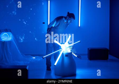 Man observing artistic installation featuring blue neon tubes arranged in a cylindrical pattern illuminated in blue-tinted room creating futuristic ambiance Stock Photo