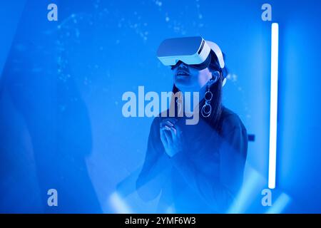 Woman immersed in virtual reality environment while using headset and accessories indoors illuminated by blue light creating futuristic atmosphere Stock Photo