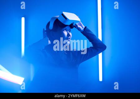 Woman experiencing virtual reality, adjusting her headset in a dimly lit room surrounded by blue lighting. Immersive technology creating a futuristic atmosphere Stock Photo