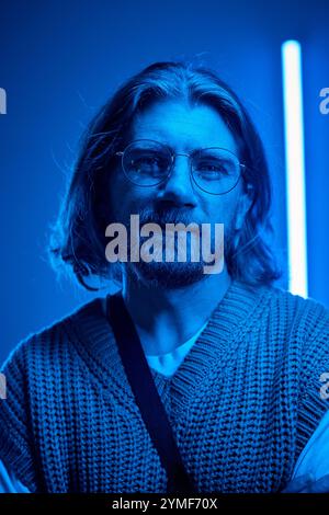 Portrait of bearded man with glasses in an atmospheric blue-toned setting, wearing a knitted sweater, presenting calm and thoughtful expression Stock Photo