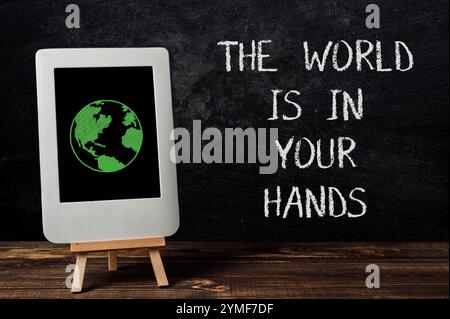 Tablet with a green globe on it is on a wooden stand. The tablet is on a black chalkboard with the words The world is in your hands written on it Stock Photo