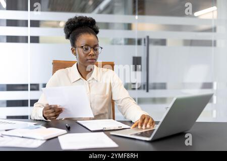 Businesswoman analyzing data on laptop in modern office holding documents. Focused professional working on project, checking reports, making notes, enhancing productivity. Stock Photo